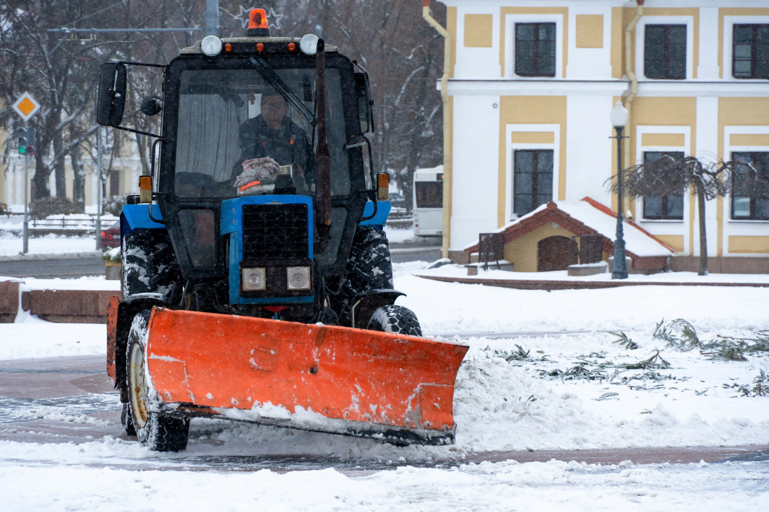 A tractor cleans snow in the city in winter after a snowfall. Cleaning city streets from snow. Tractor driver at work in the city square during a snowstorm.