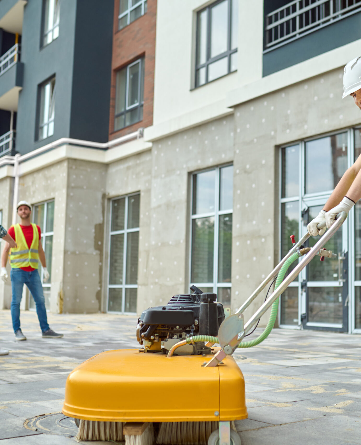 Working process. Smiling worker in protective helmet gloves and yellow tshirt cleaning construction site using special equipment and two men discussing looking at new building